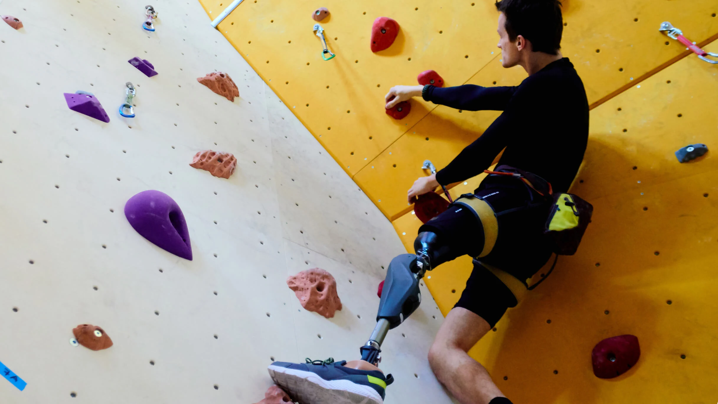 Man with prosthetic leg, dressed in black, ascends a colourful climbing wall