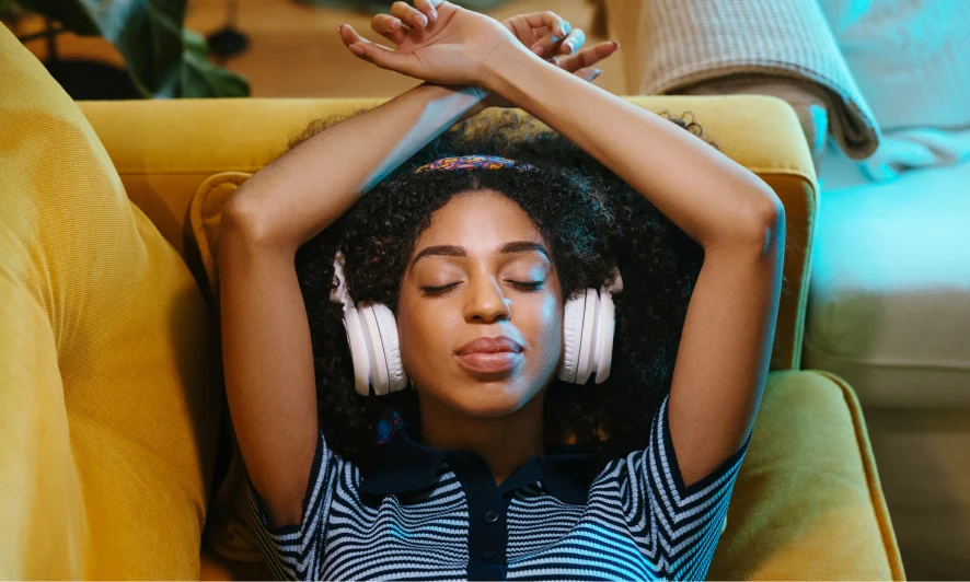 Black woman in a striped navy and light blue, short-sleeved top, eyes closed, wearing white headphones. She lies back on a soft yellow-orange couch, arms resting above her head.