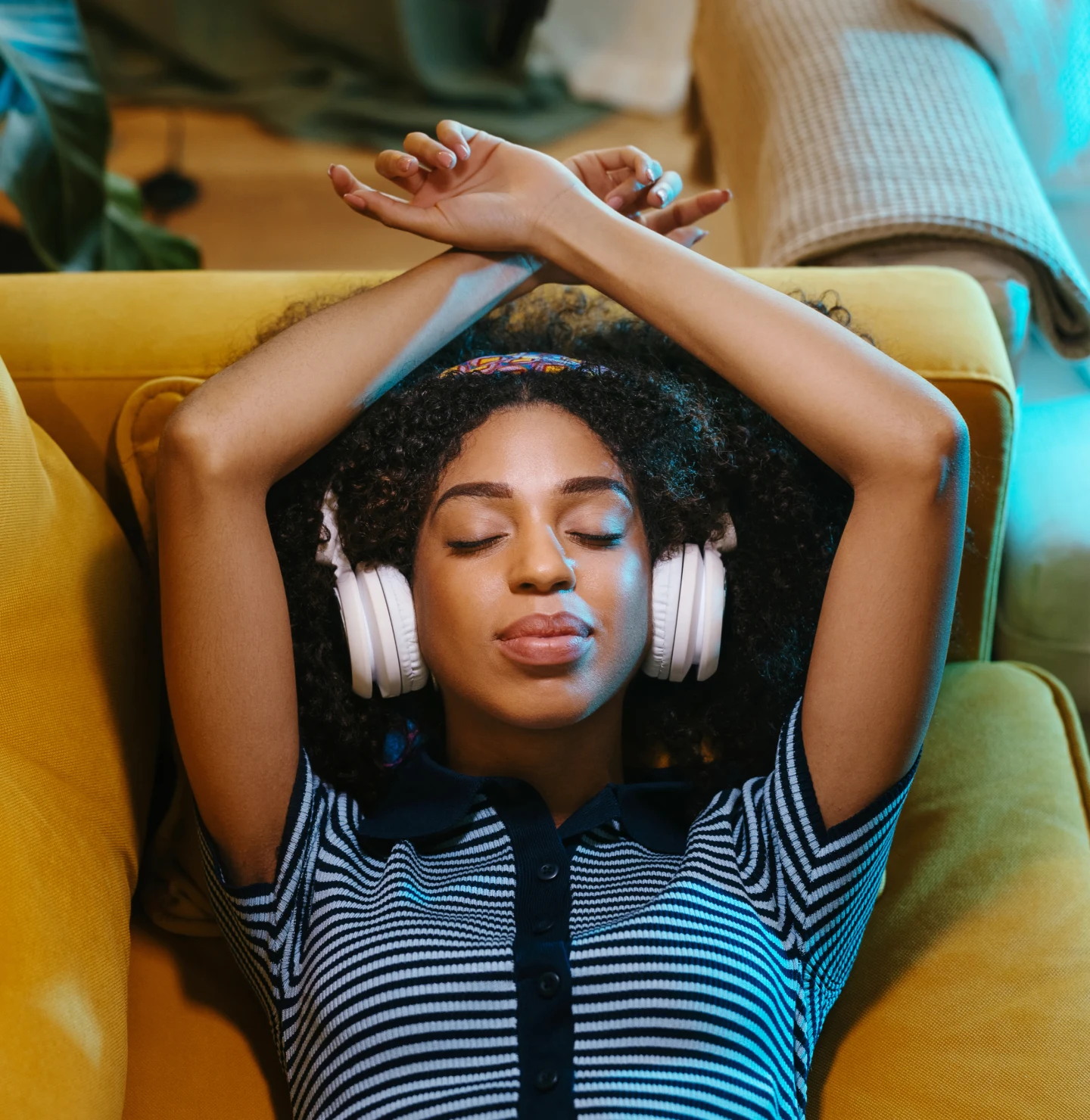 Black woman in a striped navy and light blue, short-sleeved top, eyes closed, wearing white headphones. She lies back on a soft yellow-orange couch, arms resting above her head.