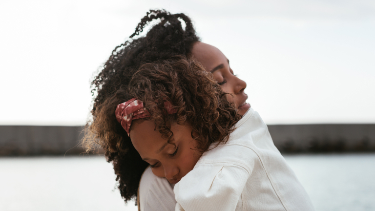 Mother and daughter wearing white dresses hugging in front of a body of water.