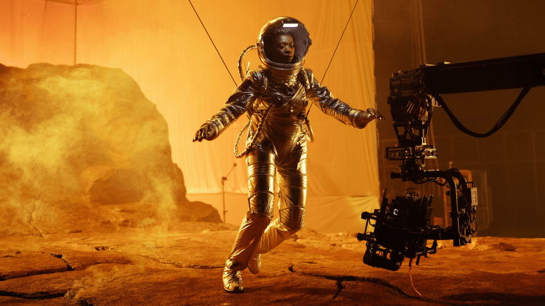 Une femme en costume d’astronaute qui pose sur un plateau, devant des roches spatiales.