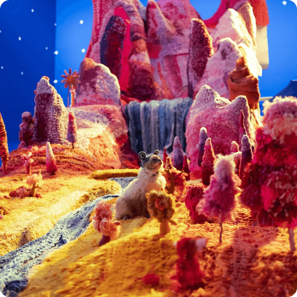 A photo of a cat wearing a crochet frog hat sitting in a whimsical crochet landscape.