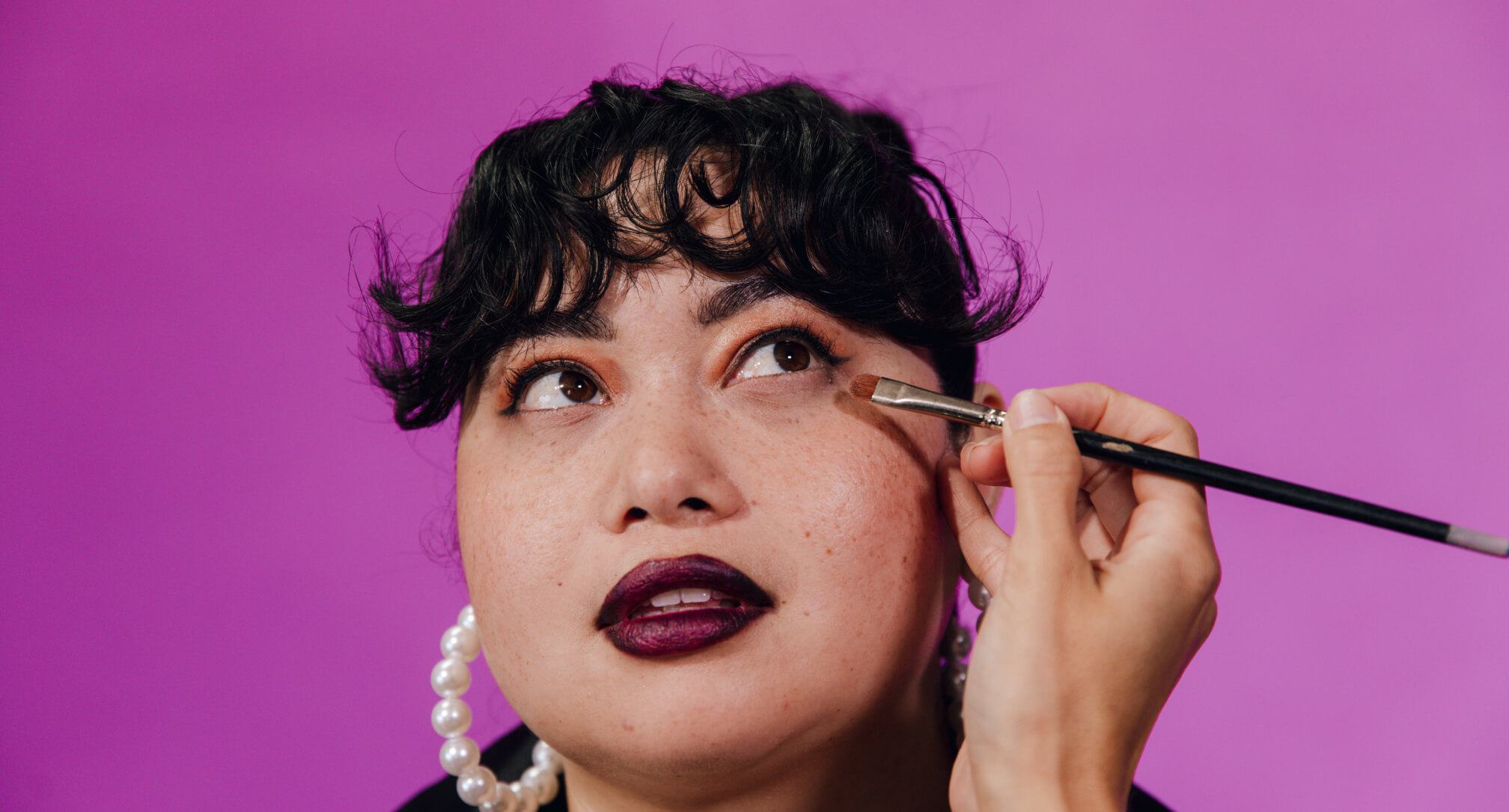 Dark-haired woman with curly bangs looks up and to the side as someone applies her eye makeup