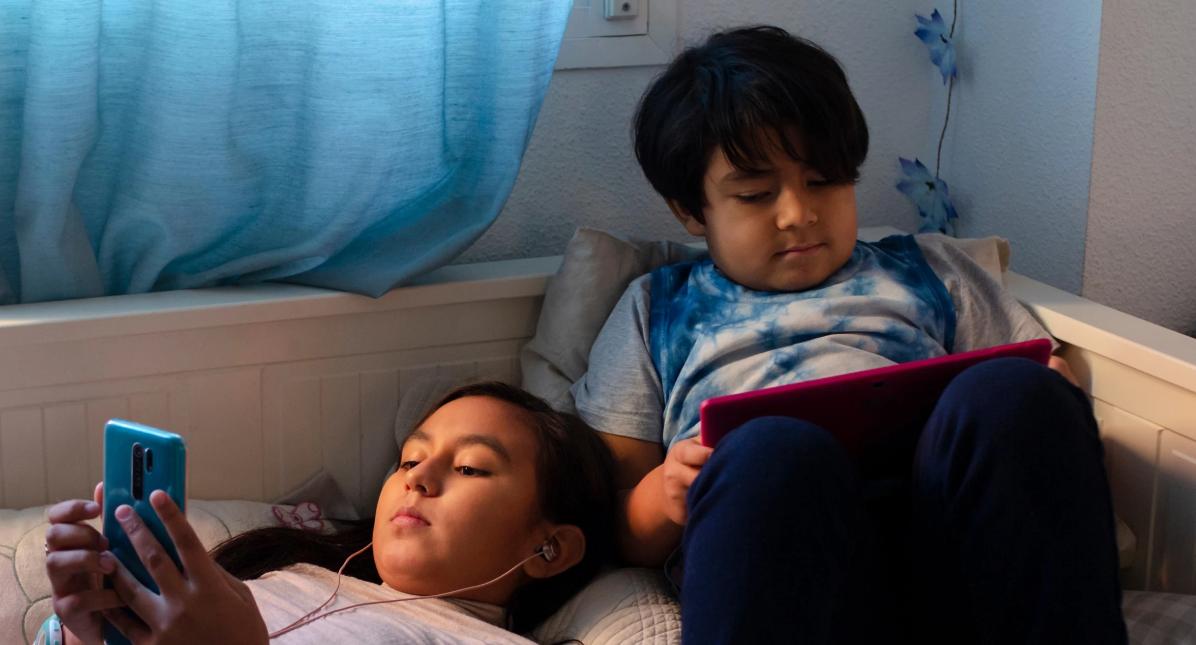 A Latinx girl and boy lounge on a bed with their mobile devices in hand