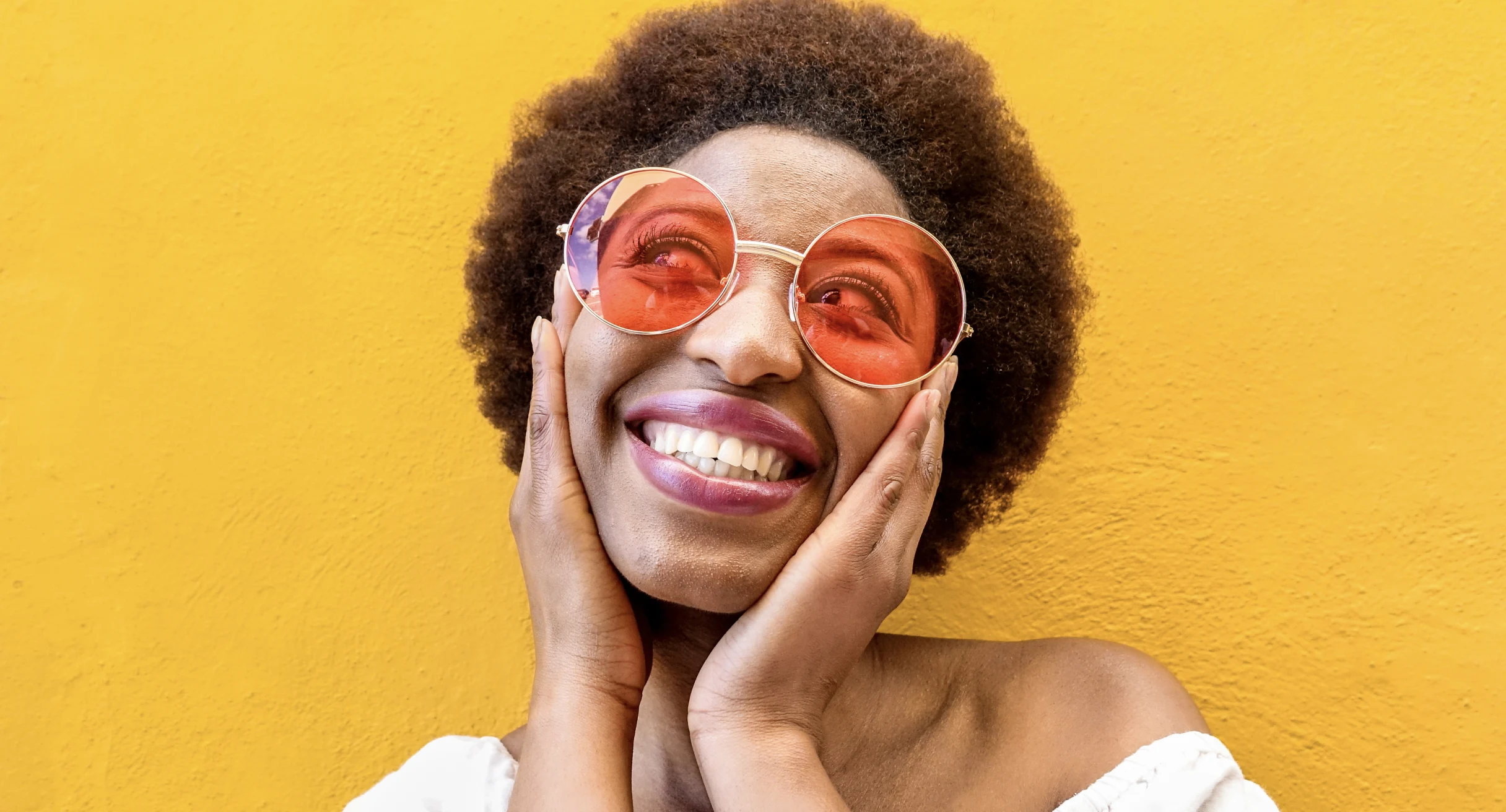 A Black woman in large, tinted glasses smiles and holds her cheeks in front of a yellow wall 