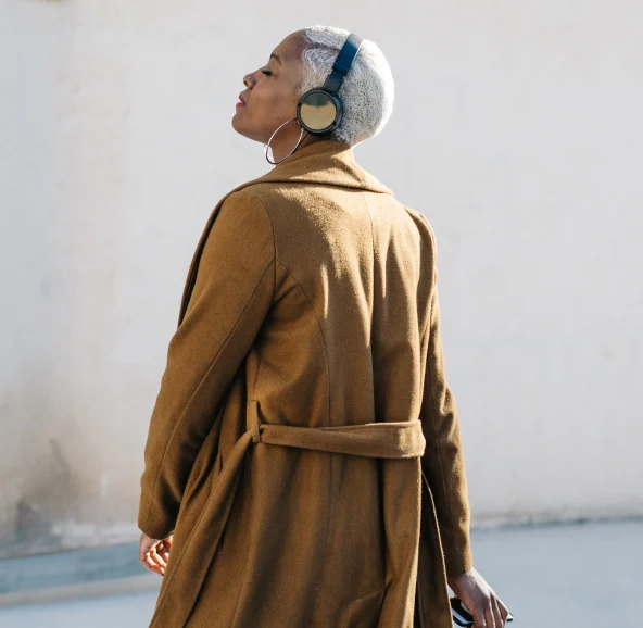 A Black woman with short white hair in a Brown trenchcoat and wireless headphones