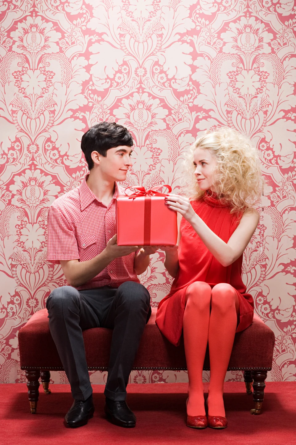 Homme blanc avec des cheveux courts foncés qui porte une chemise rose et un pantalon foncé et femme blanche en robe rouge avec des cheveux blonds épais, assis sur un banc rouge, l'un en face de l'autre, avec un papier peint à motifs roses en arrière-plan. Ils tiennent ensemble un cadeau dans un emballage rose avec un nœud rouge.