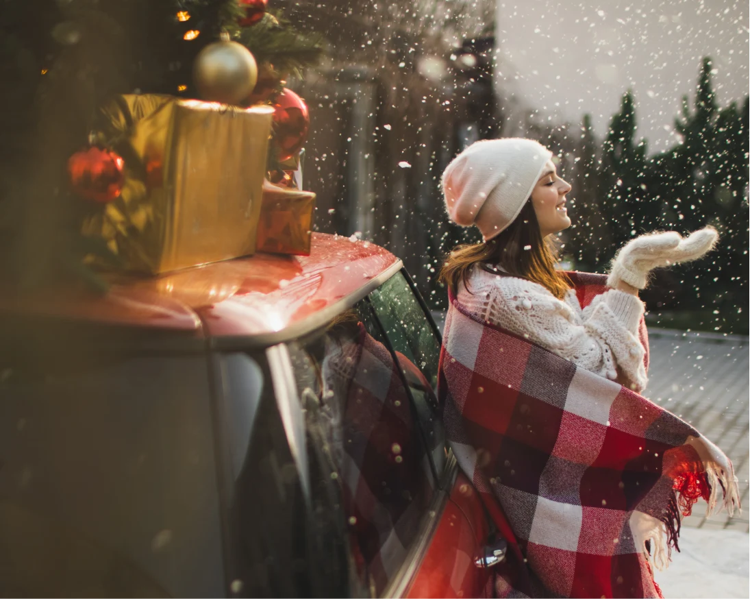 雪の中で、白いニット帽と手袋、ニットを着た女性が、クリスマスツリーとプレゼントを乗せた赤い車に寄りかかっている様子。