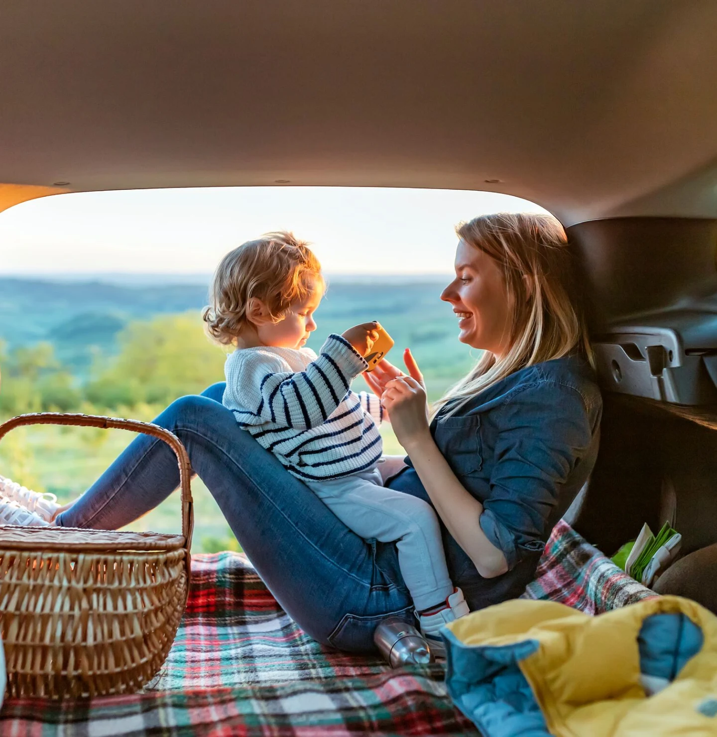 A mother and her child sit in the back seat of their car having a picnic in the countryside.