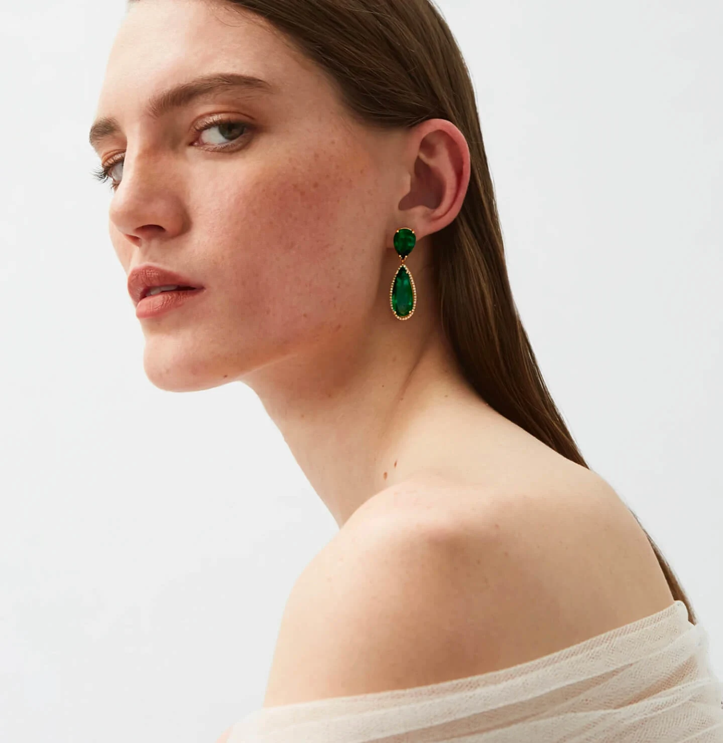 White woman with freckles and long brown hair, wearing a white off-the-shoulder tulle top and green earrings, gazes into the camera 