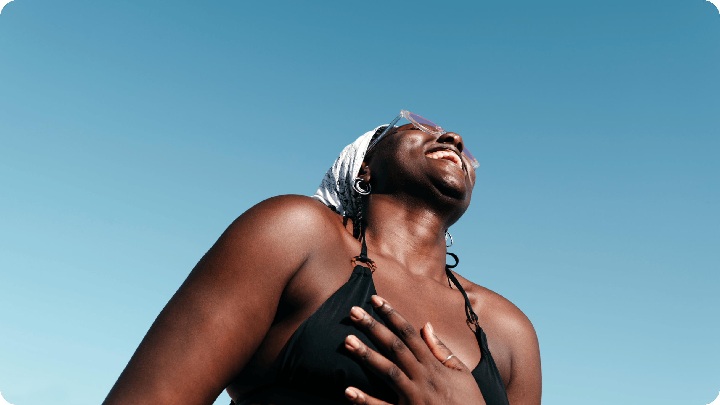 A woman in a bathing suit and sunglasses, smiling and looking up at a bright blue sky.