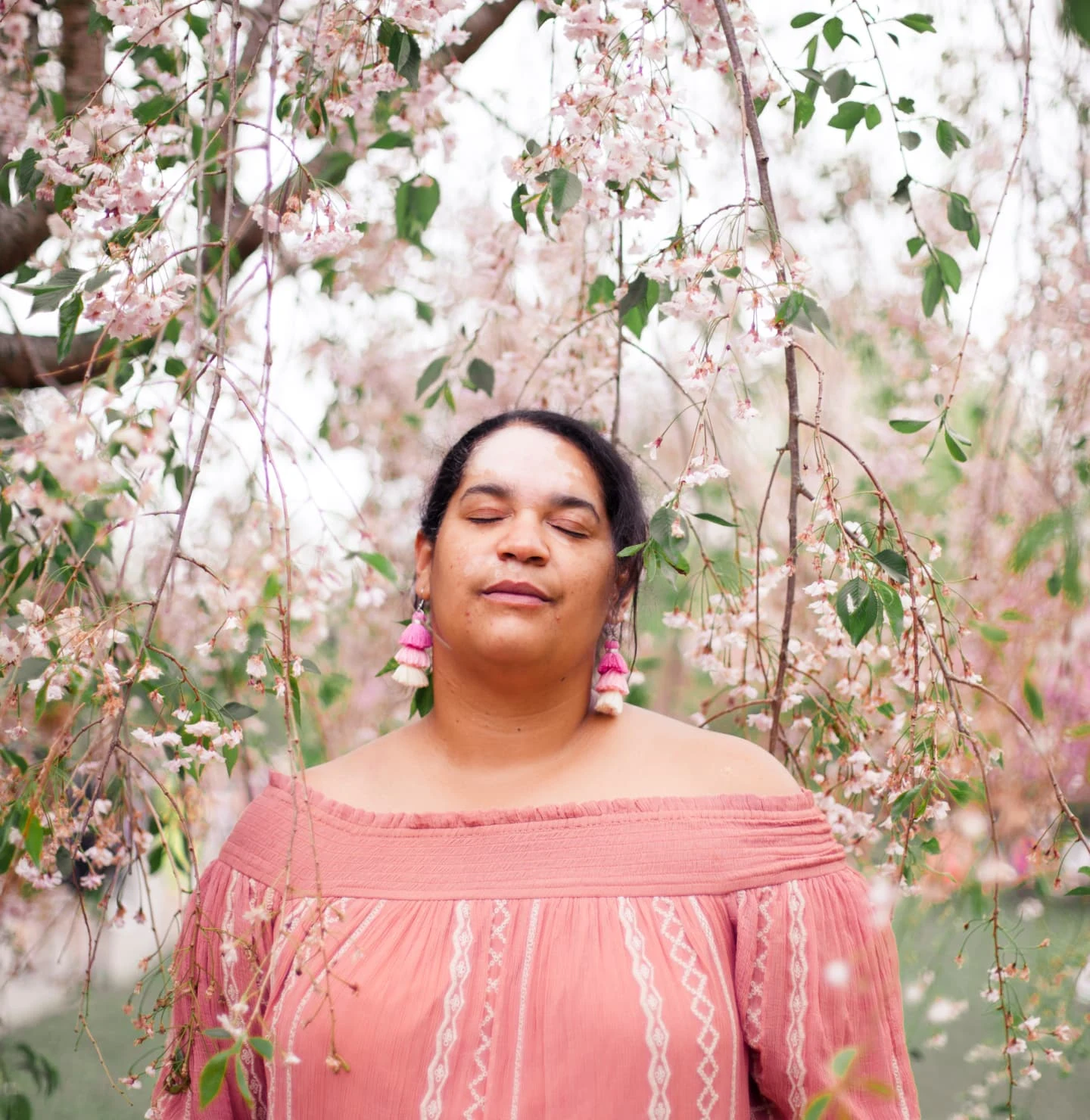 A Black woman in pink stands with her eyes closed under the white and pink blossoms of a tree