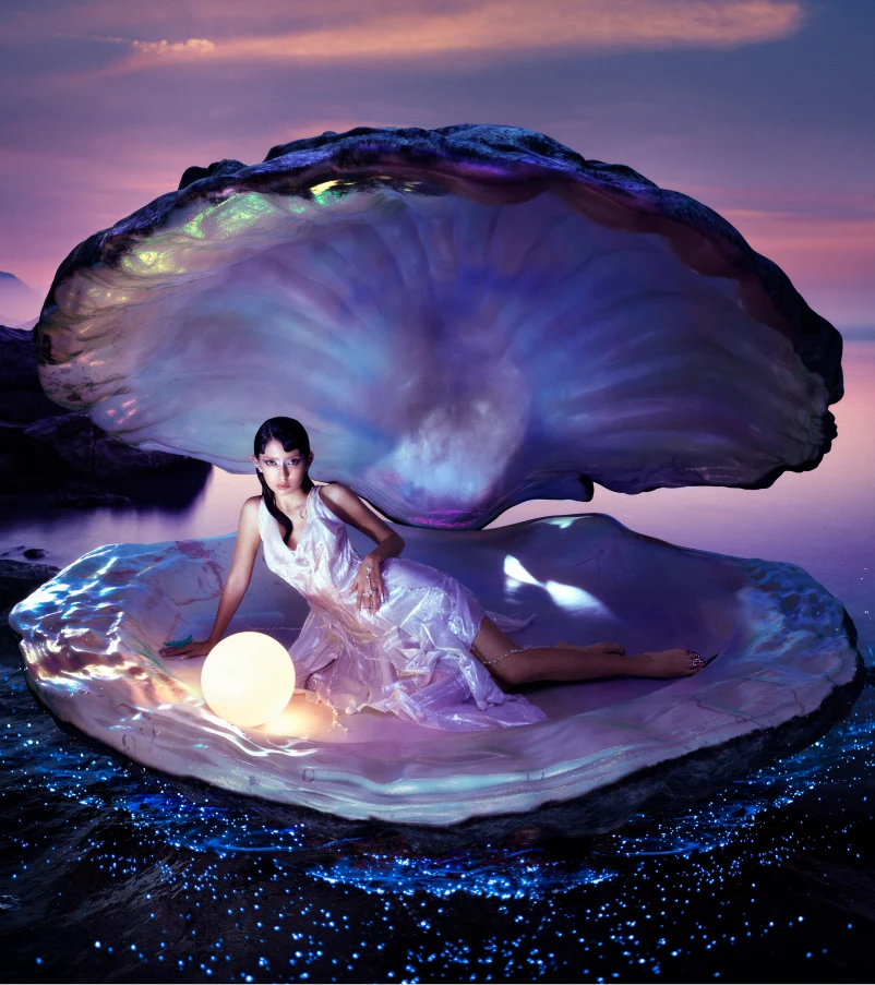 A woman in a wet-looking dress reclining inside a giant, pearlescent clam shell with a glowing pearl in it