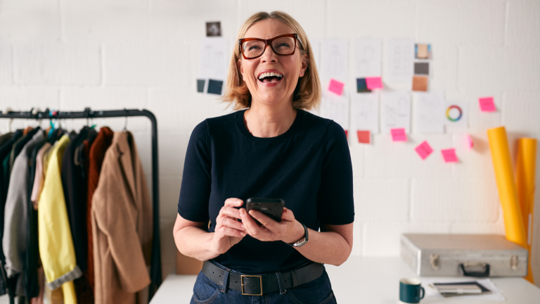 Older white woman with blonde hair and glasses looking up from her smartphone and laughing