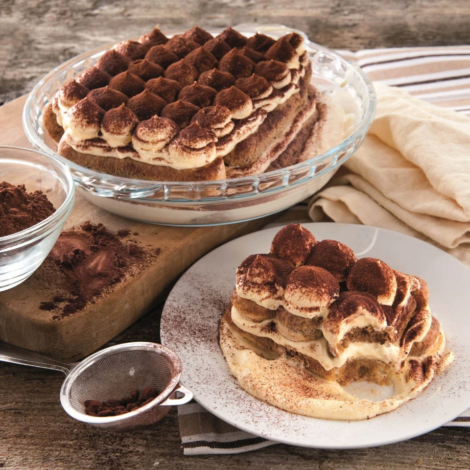 Tiramisu servi dans une grande verrerie ronde à côté de la poudre de cacao et du plumeau, avec une tranche servie dans une assiette séparée et une bouchée prise