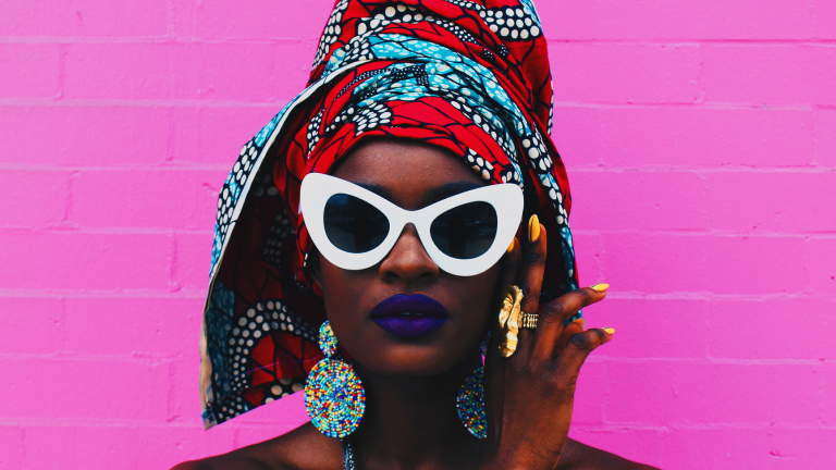Ein Foto zeigt eine schwarze Frau von den Schultern aufwärts vor einer leuchtend rosa Backsteinwand. Die Frau trägt lila Lippenstift, eine weiße Sonnenbrille, ein rot-blaues Kopftuch und klobigen Schmuck.