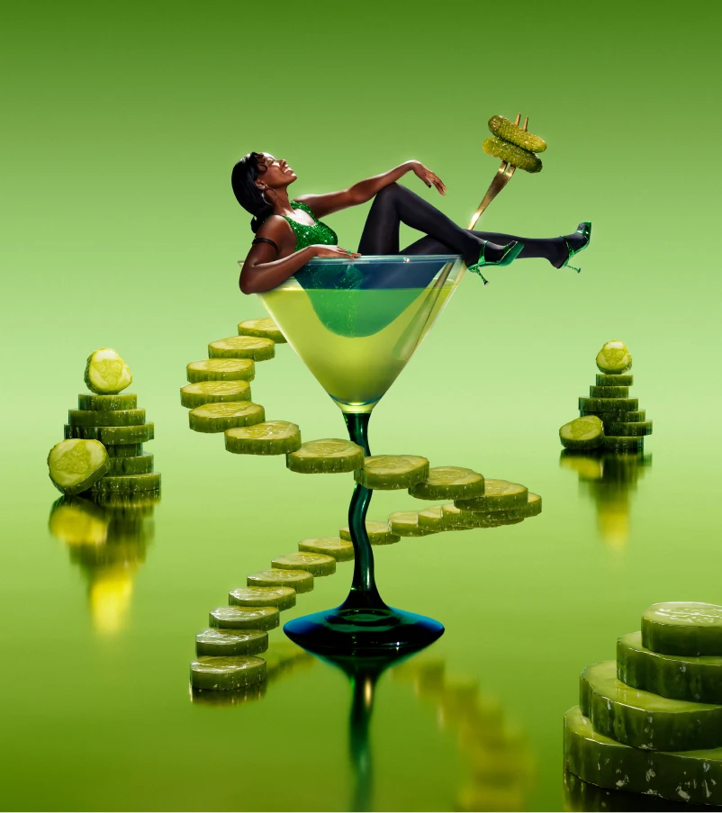 A woman in a green sparkly dress, black tights and metallic green strappy heels sits in an emerald green martini glass. The glass has a long dark green stem and sits against a bluish-green background.