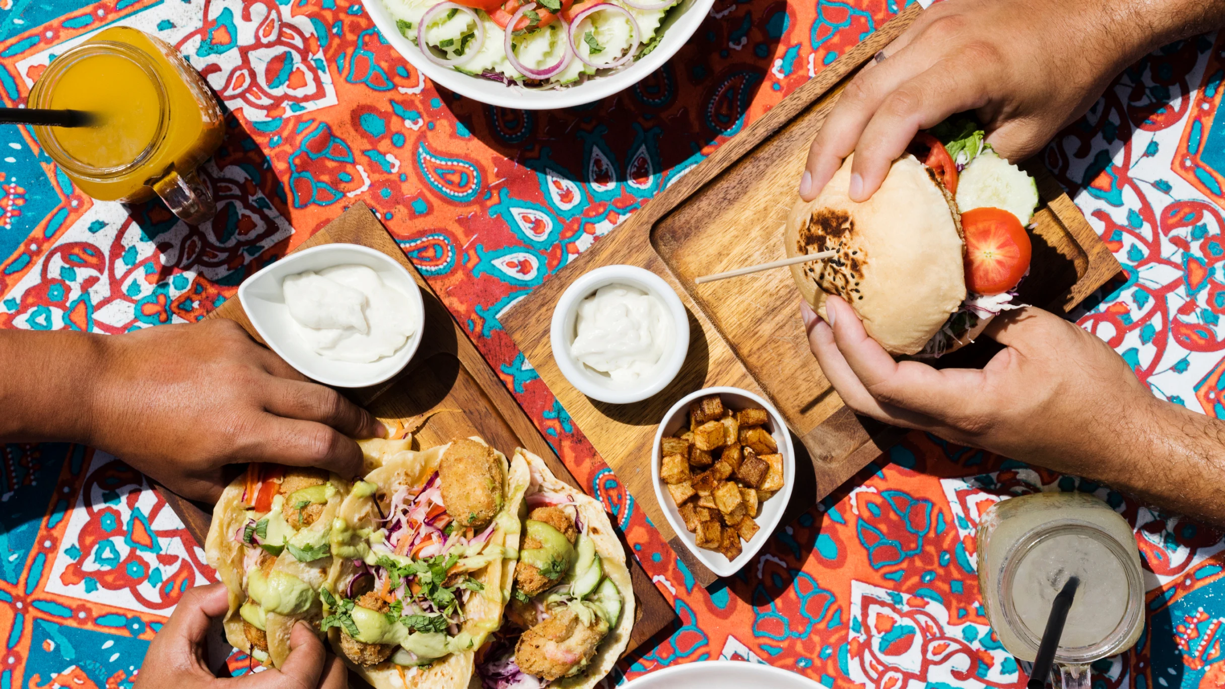 Aerial view of food items on a colorful patterned table cloth: lemonade and orange juice in glass jars, white hands holding a veggie burger, spaghetti and meatballs, brown hands holding one of three fried fish tacos, a falafel bowl.