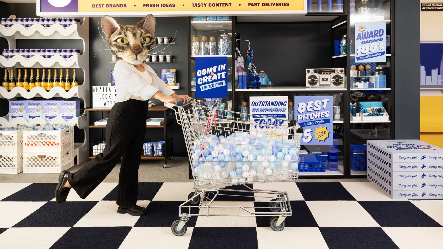 Person wearing a giant cat head pushing a shopping cart through a bodega-inspired boardroom.