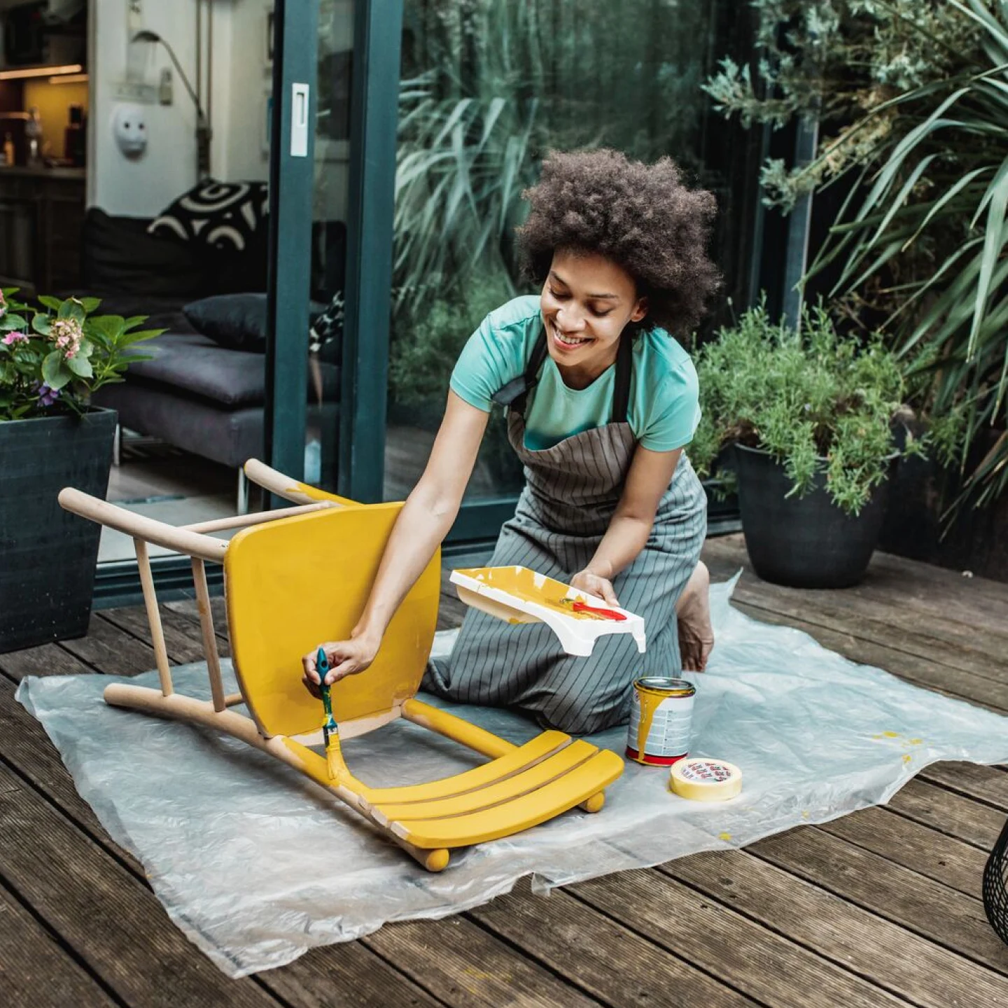  a smiling Black woman in coveralls paints a yellow wooden chair on a wooden backyard patio. Green plants in the on right. An open sliding door on the left, living room with dark gray couch inside.