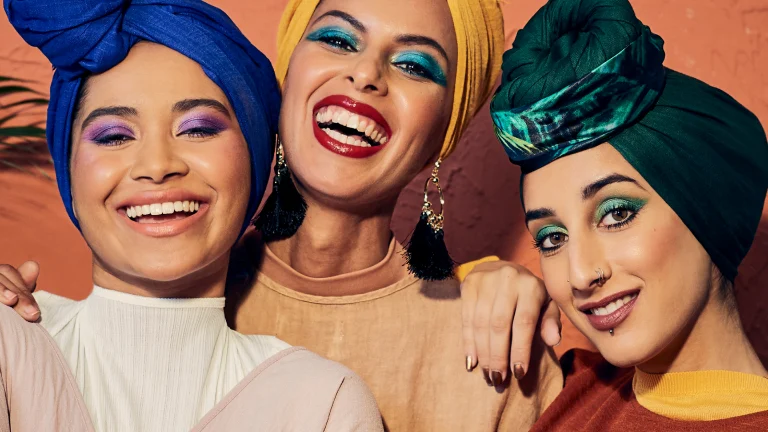 Three smiling women, each dressed in a head scarf of a different color: deep blue, bright yellow and deep green