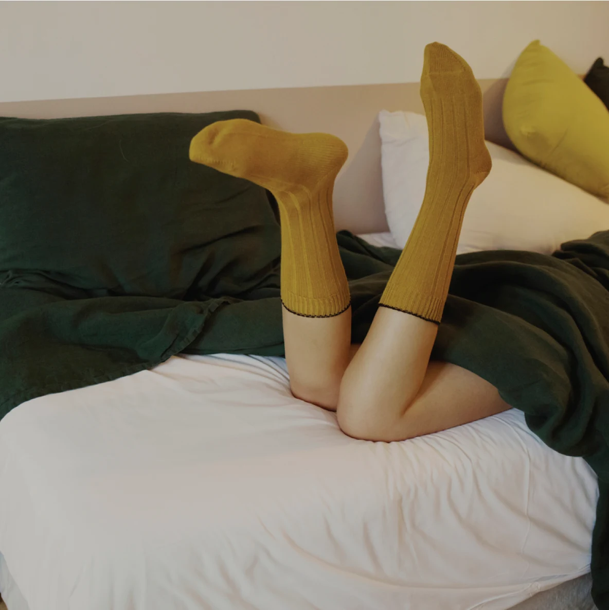 A shot of a woman’s legs wearing mustard colored socks on a bed with assorted pillows covered in a green blanket.