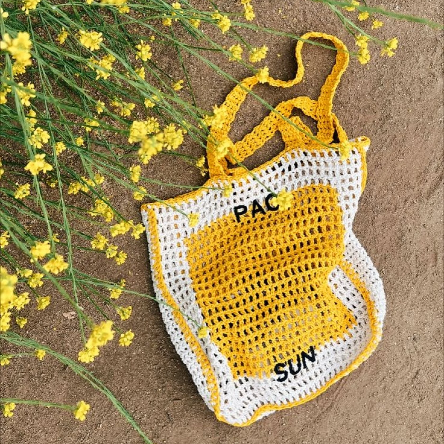 A crocheted white and yellow bag that reads Pac Sun lies on the ground next to yellow wildflowers