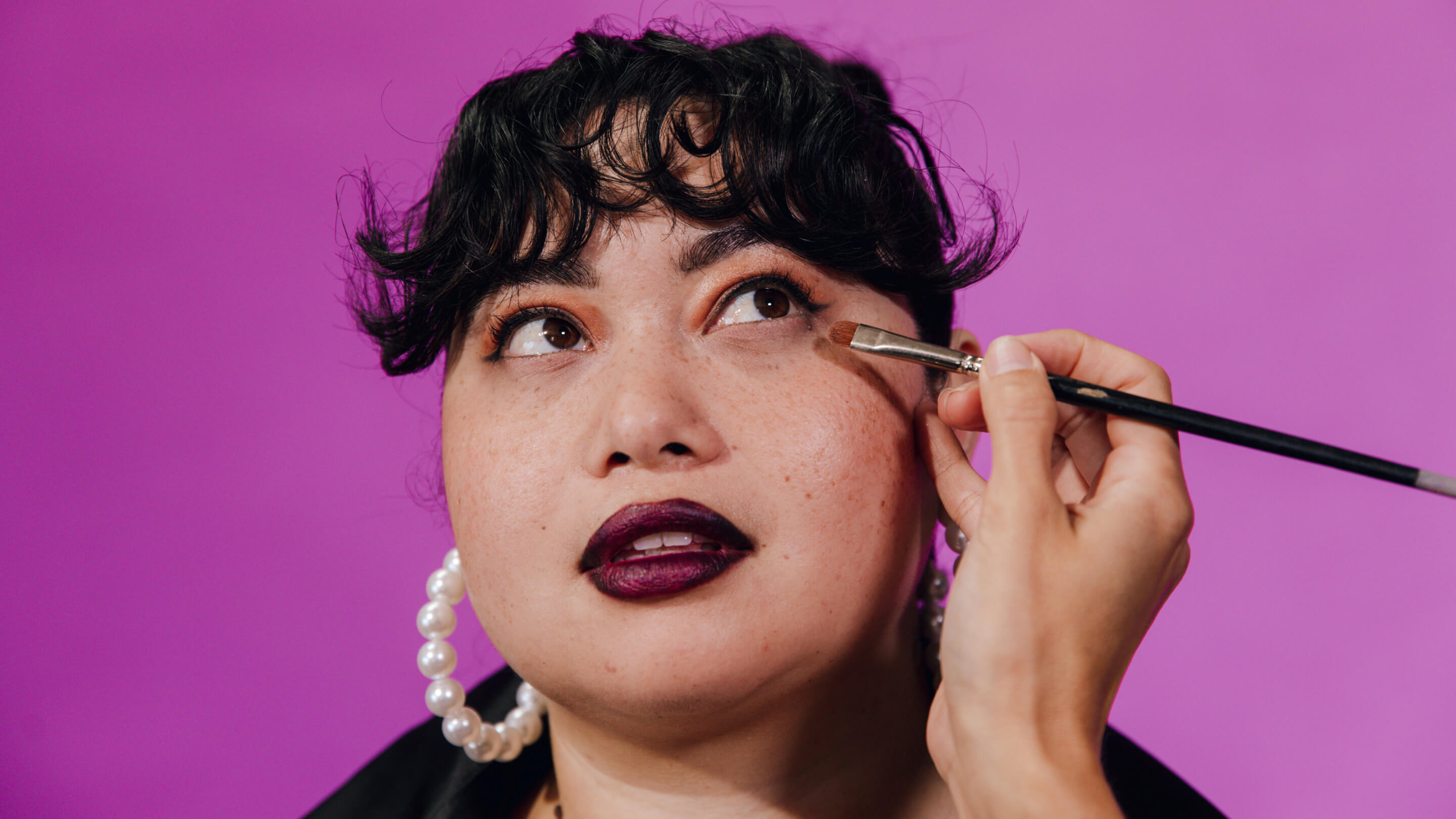 Dark-haired woman with curly bangs looks up and to the side as someone applies her eye makeup