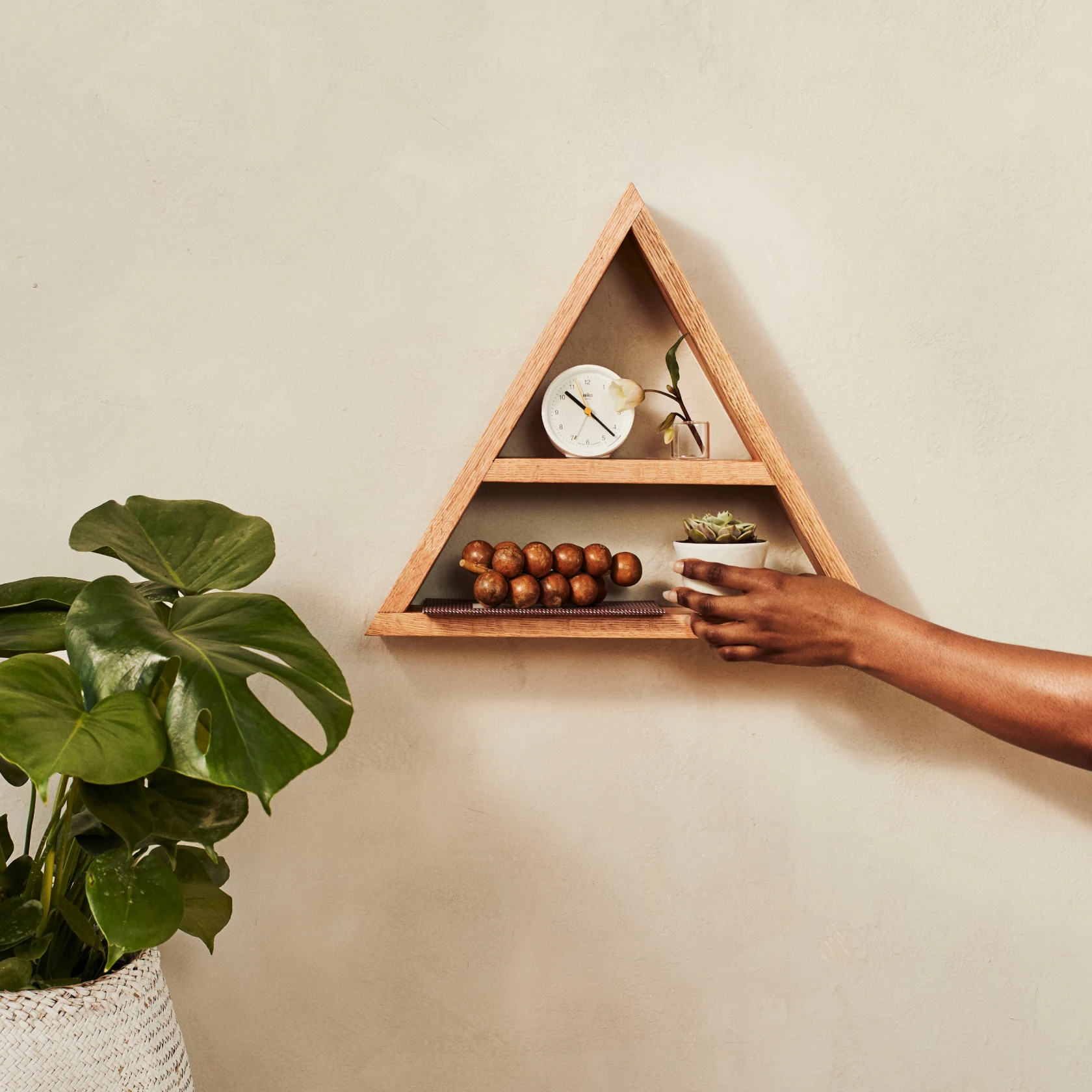 A person placing a small pot of succulents onto a floating wooden, triangle shelf hanging on a plain white wall. On the shelf is also a wooden clock, one white flower in a small glass and wooden sculpture of grapes. A large plant is on the floor next to the shelf.