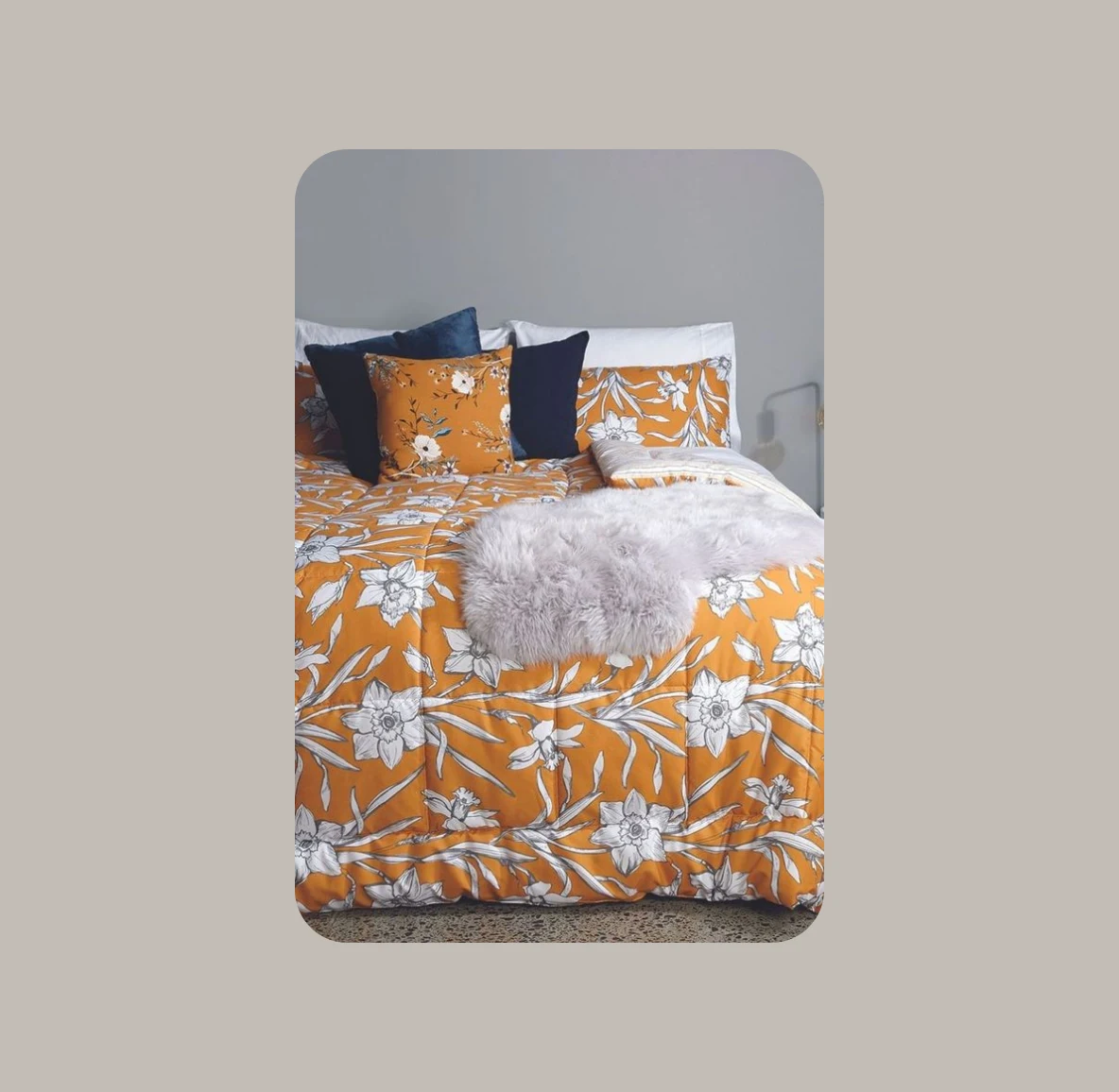 Pictured is a bed with a patterned comforter. The comforter is a mix of orange, navy and white colors with a shag white blanket laid on top of the bed. 