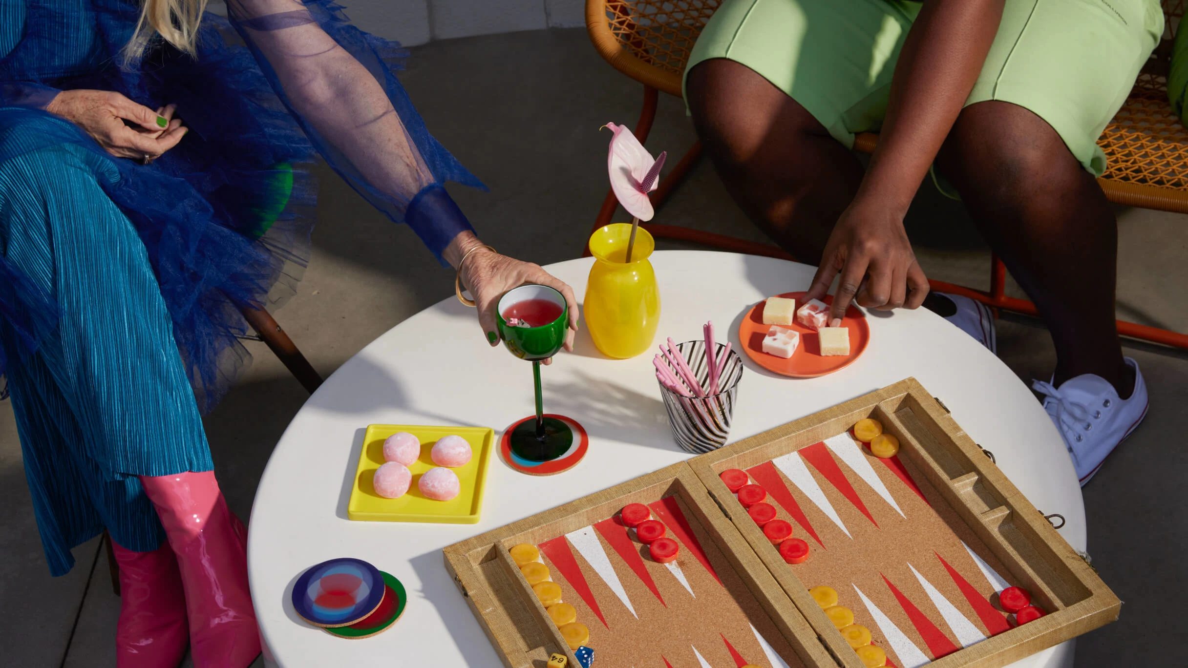 Two friends dressed in colourful outfits sit around a low white outside table playing a board game, drinking fruity drinks and eating delicious snacks. A flower in a yellow vase is on the table while pink paper straws sit in a black and white striped cup.