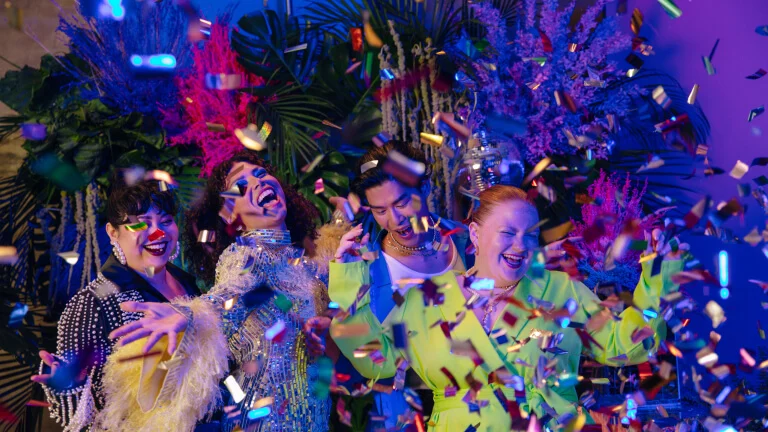 Un grupo de jóvenes vestidos con ropa de fiesta colorida y brillante, sonriendo y bailando mientras cae confeti a su alrededor.