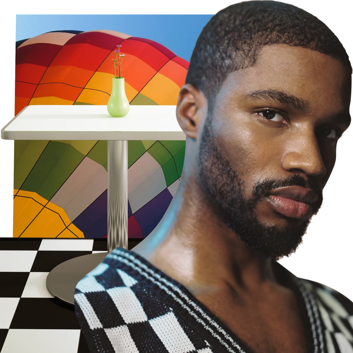 Ein Schwarzer Mann mit kurzem Haar in einem Strickpullover mit Schachbrettmuster und V-Ausschnitt blickt in die Kamera. Ein weißer Tisch, auf dem eine schmale hellgrüne Vase steht. Im Hintergrund ist ein Heißluftballon in Regenbogenfarben zu sehen.