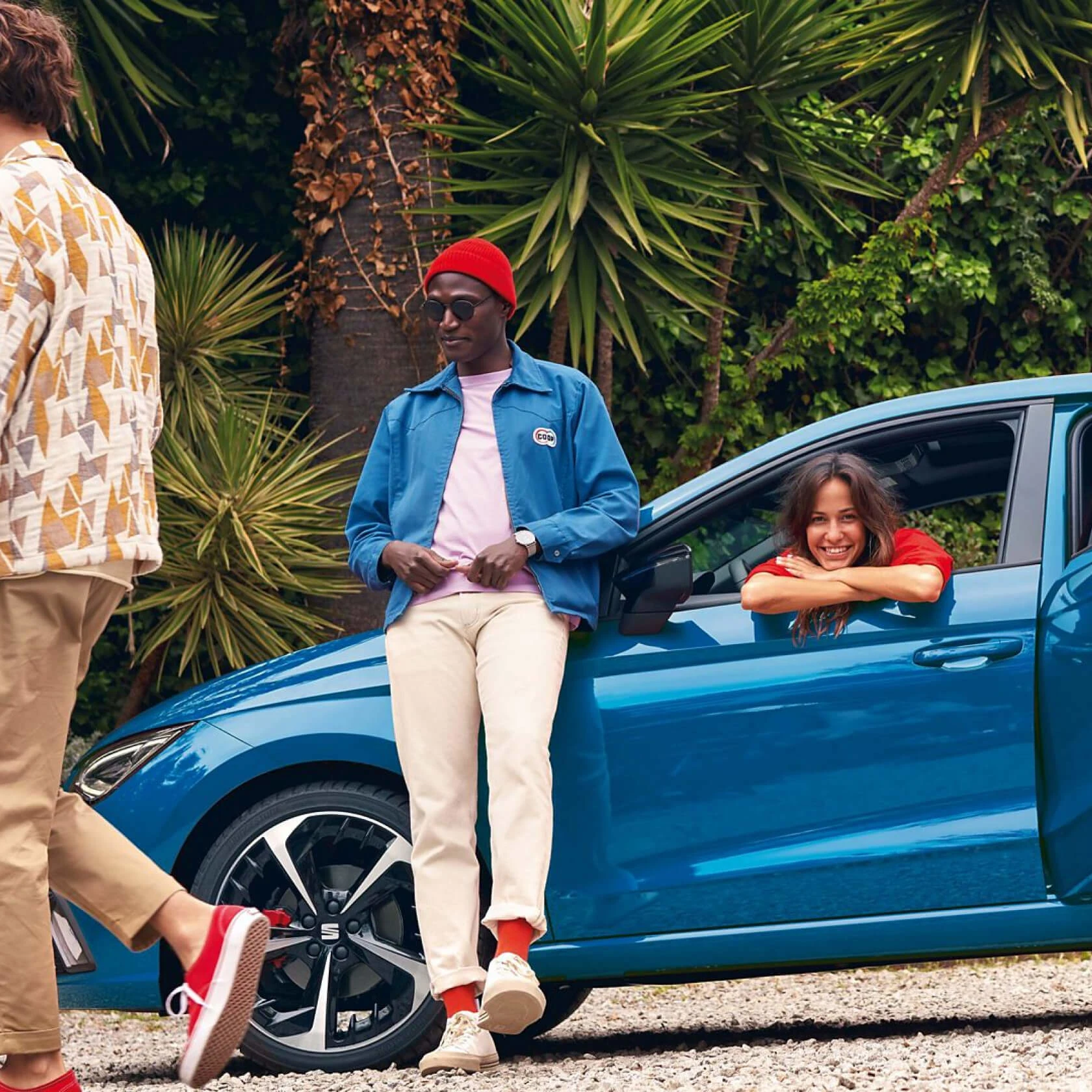 Stilvoller Mann lehnt an blauer Limousine, die draußen neben grünen Palmen geparkt ist, und junge Frau lächelt aus dem Fahrerfenster