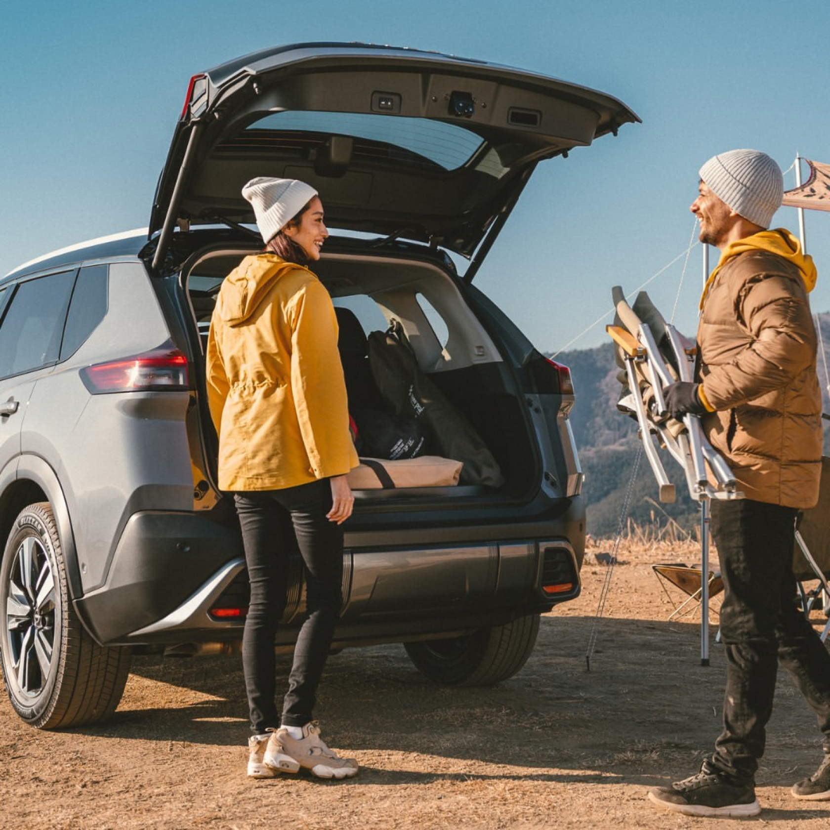 Una pareja joven vestida con parkas y gorros en un área al aire libre, empacando equipo de campamento en el maletero de una camioneta