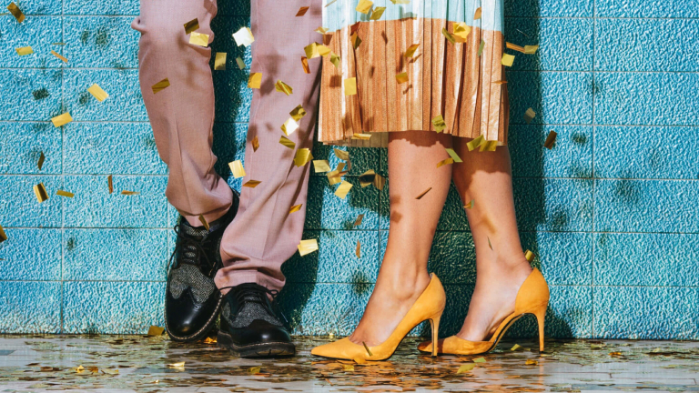Pernas de duas pessoas próximas uma da outra contra uma parede azul, cercadas por confetes. A pessoa à esquerda veste uma calça na cor salmão e sapato social preto. A pessoa da direita usa uma saia bicolor e sapato de salto amarelo.