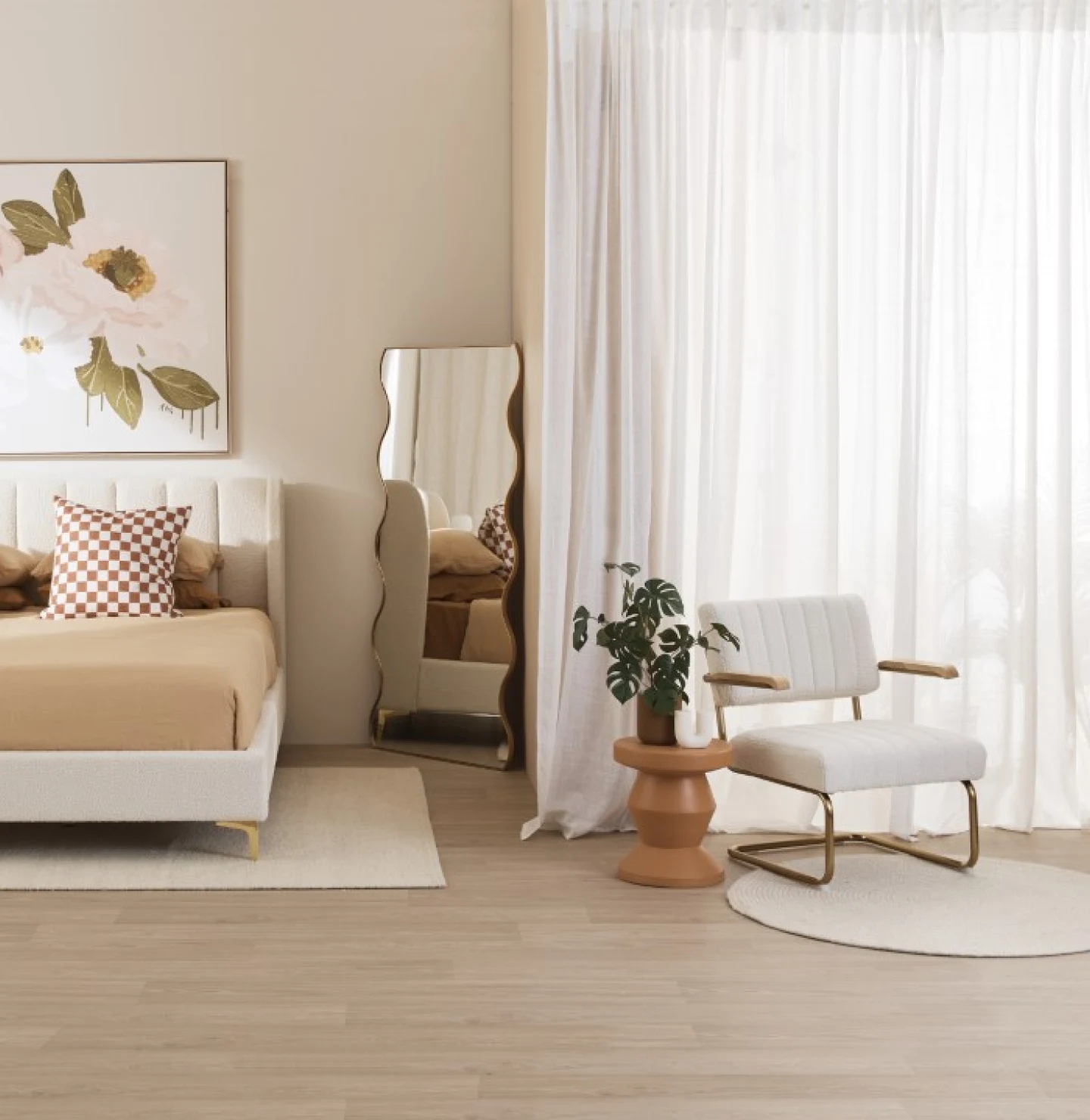 An image of a bedroom painted with tan undertones. To the left, a brownish tan bed with a checkered pillow and a floral painting above it with a curvy mirror in the center. To the right, a white and gold metal chair sitting next to a plant. 