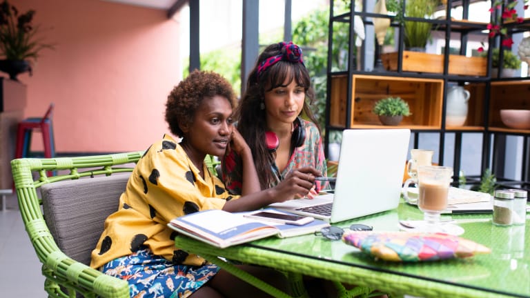 Two young women looking at a laptop surrounded by books and coffee.