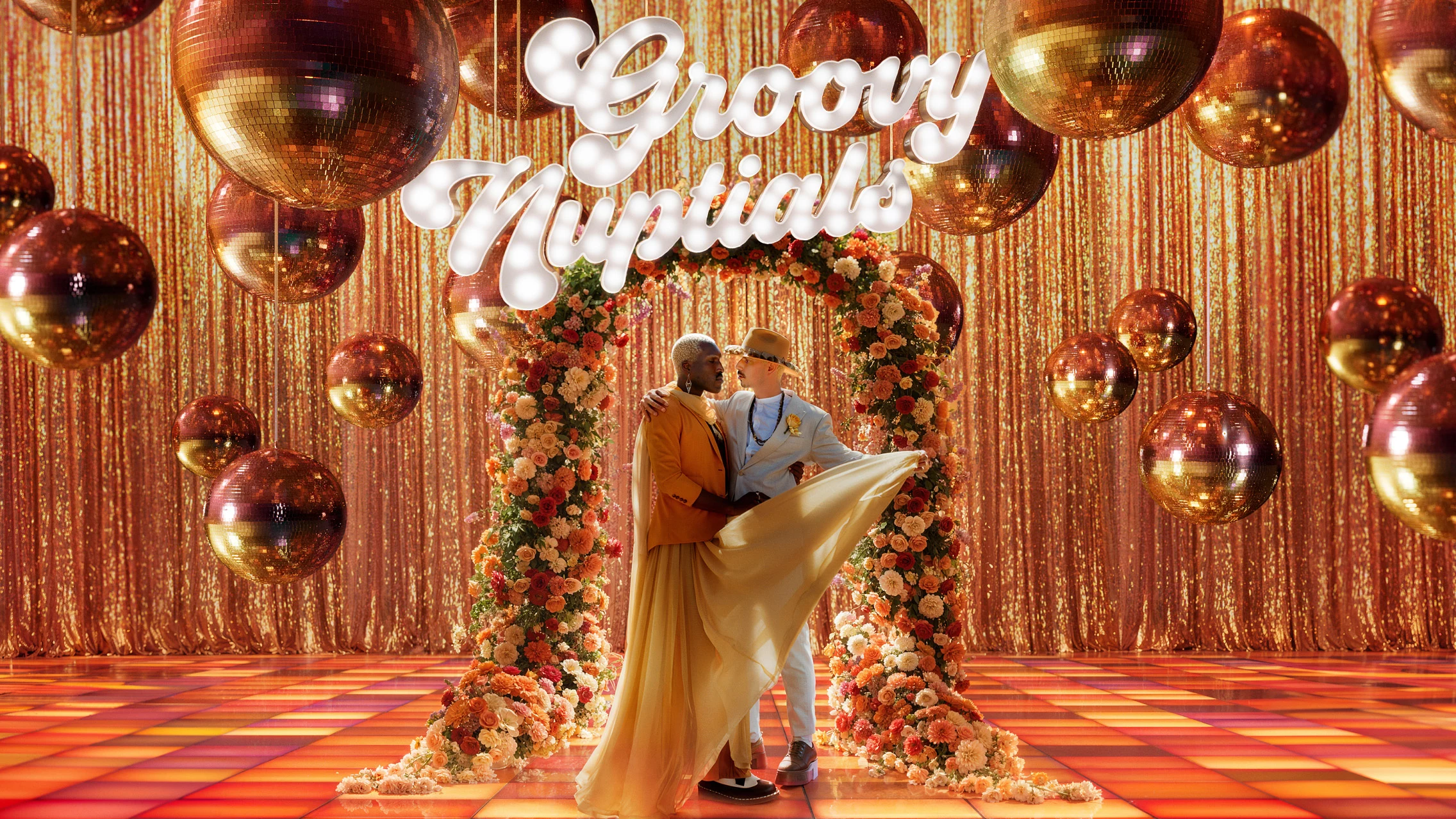 Male couple embracing under a floral arch standing atop a colorful tiled floor with sparkly rust-colored disco balls overhead. A white neon sign reads “Groovy nuptials” above their heads in cursive font.