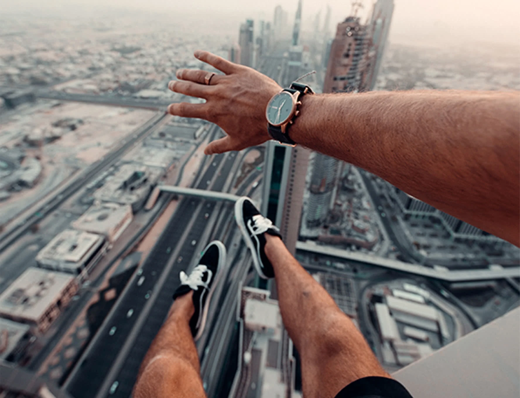 Nohy obuté v černých teniskách visící ve vzduchu a ruka s hodinkami nad rozlehlým městským panoramatem