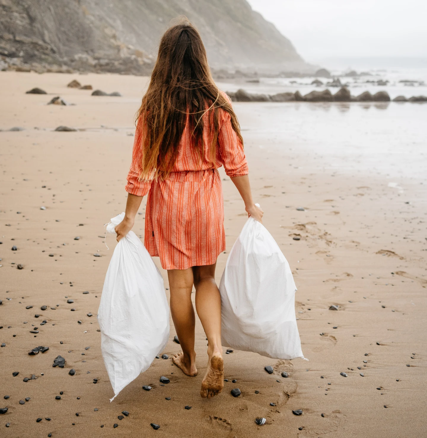 A woman in an orange dress walks barefoot on a secluded beach, carrying two, full white trash bags