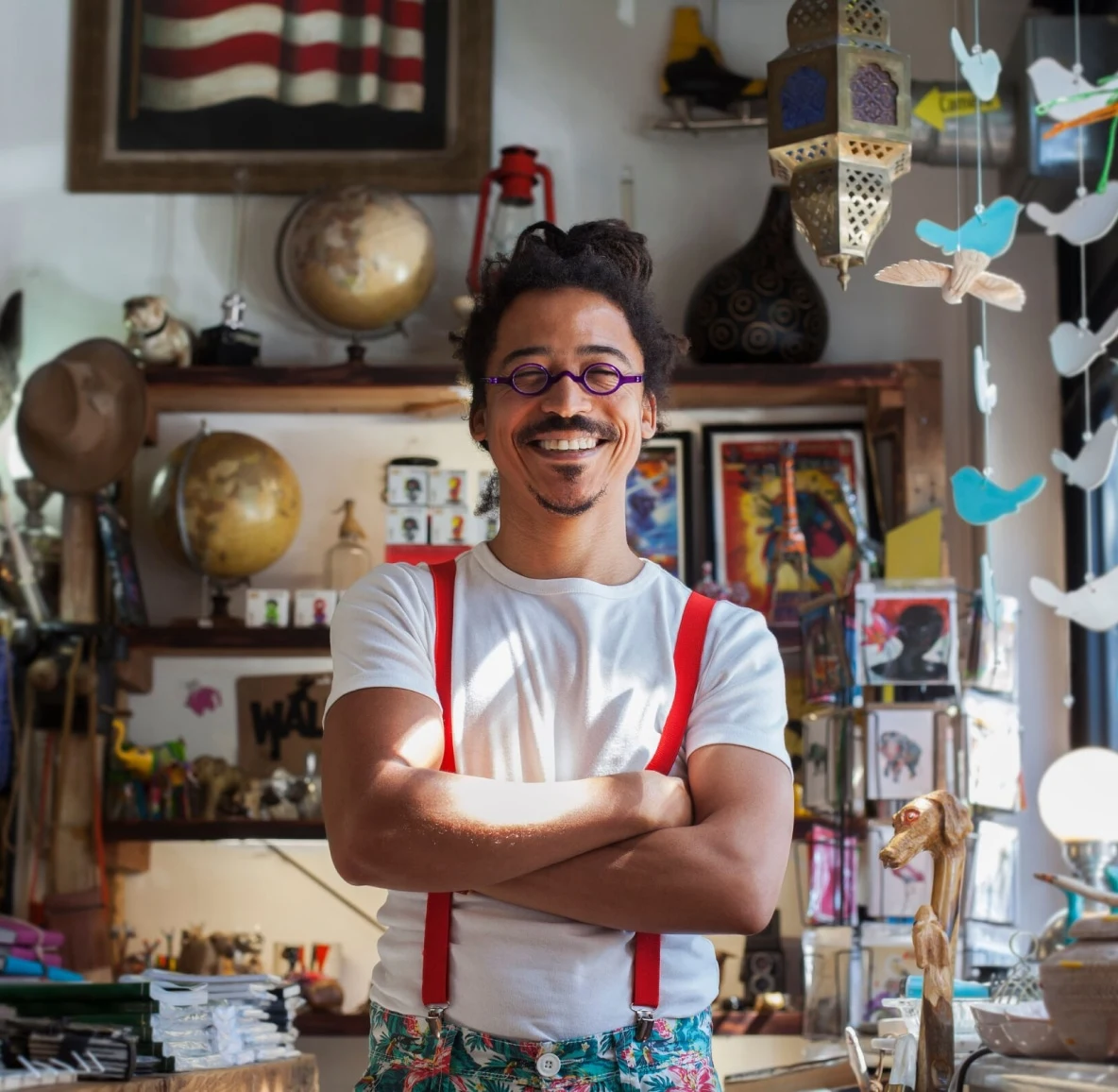 A smiling Black man with locs and red suspenders stands in a curio shop, arms crossed