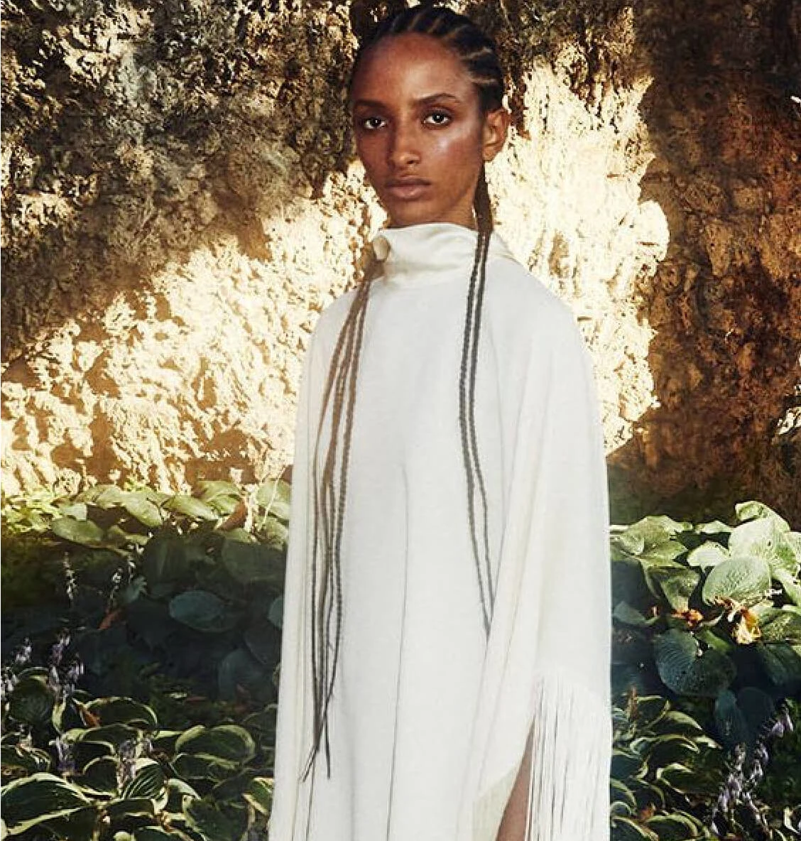Black woman with braids stands outside in front of green vegetation and a cave, wearing a white dress with turtle neck and fringe.