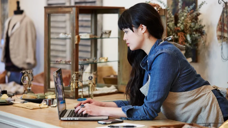 East Asian woman standing at a laptop that’s on a wood shop counter. Store in the background with a glass cabinet and suit display. 