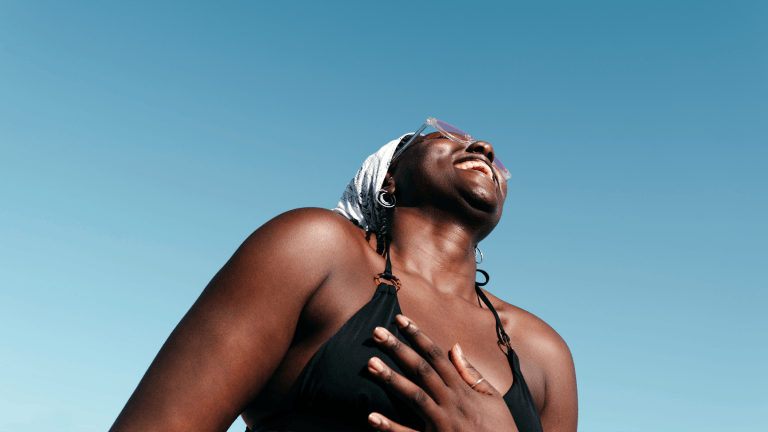 A woman in a bathing suit and sunglasses, smiling and looking up at a bright blue sky.