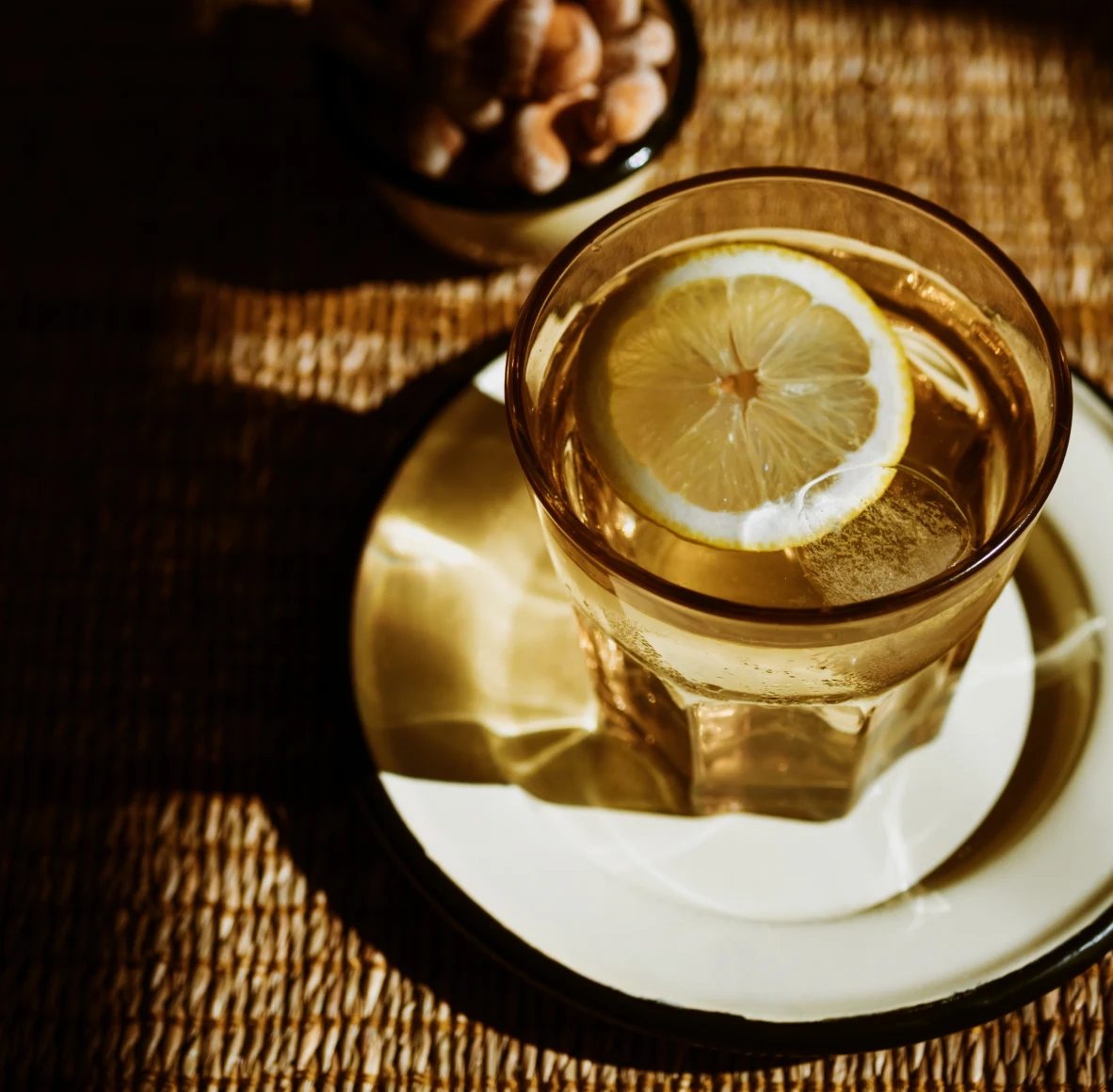 A glass of water with lemon on top of a small white plate with a bowl of nuts behind it slightly out of focus
