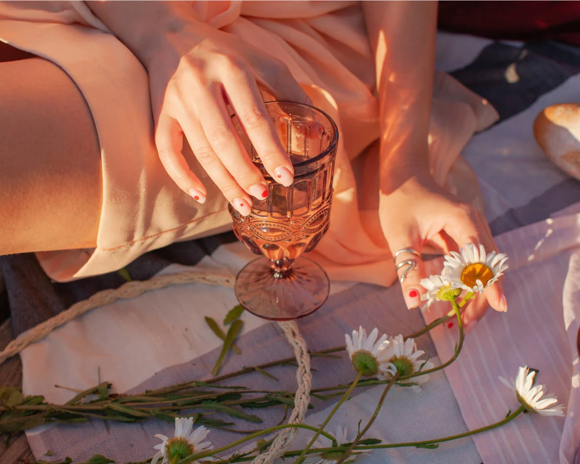 Woman with painted nails and a pink dress sits on a picnic blanket holding a glass of wine and picking daisies.