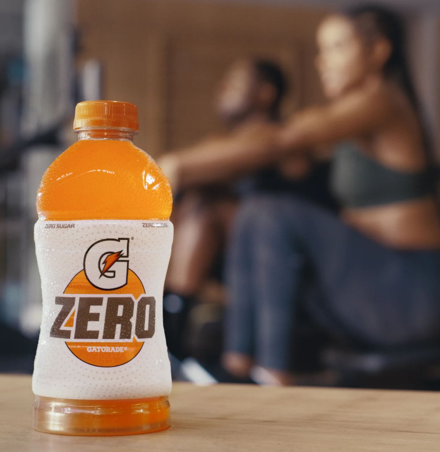 In the foreground, a bottle of orange-flavoured Gatorade Zero. In the background, two people in workout clothes are on rowing machines.  