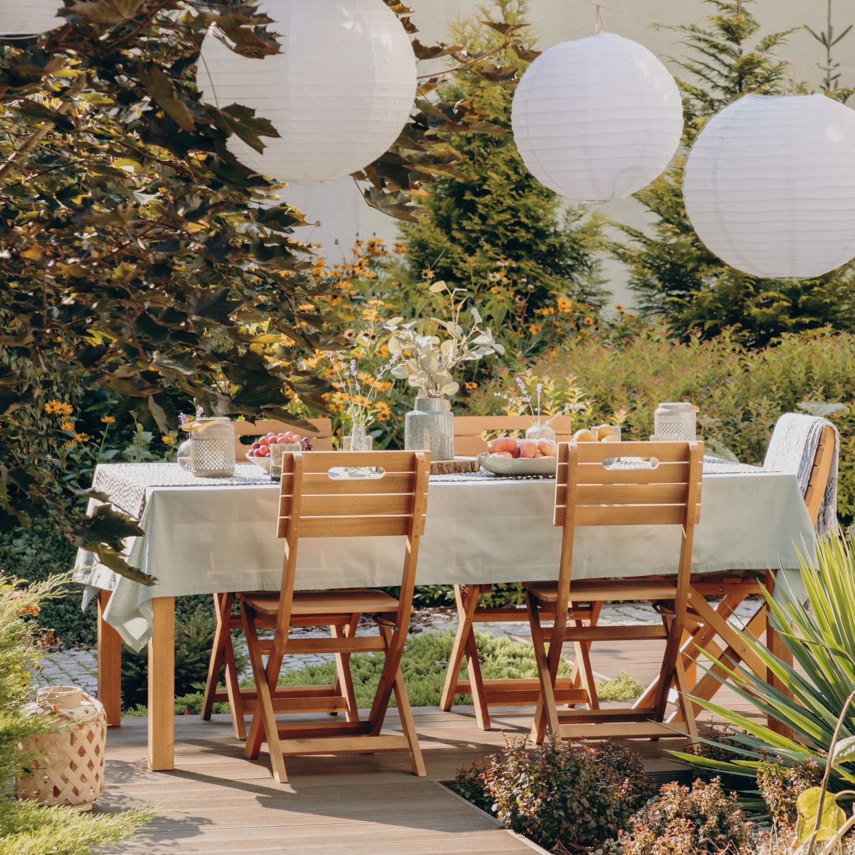 Outdoor patio with wooden table styled with tablecloth, flowers, fresh fruit, and hanging paper lantern lights above