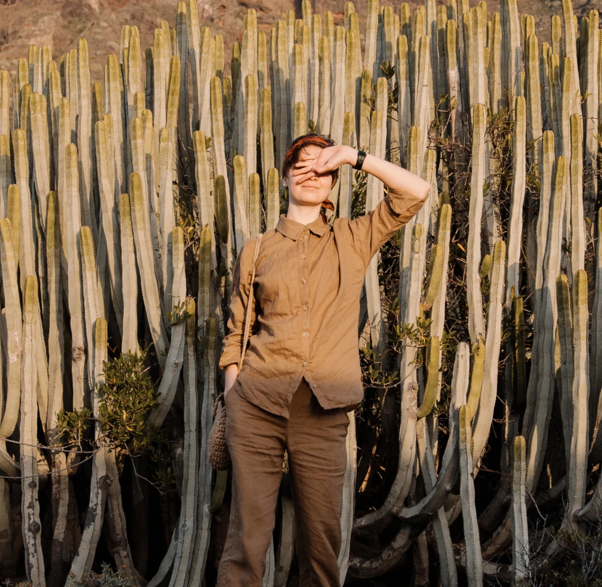 Una mujer blanca con un traje beige casual, protegiendo sus ojos del sol frente a finas plantas de cactus